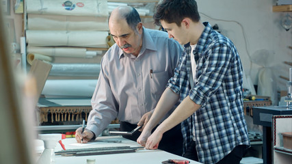 Young trainee studying how to construct a frame, senior worker talking to him behind the desk in frame workshop