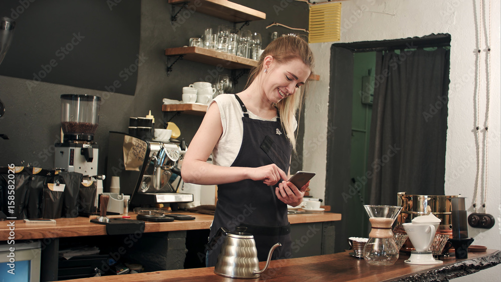 Poster cheerful and content female barista using mobile phone and texting at coffee shop