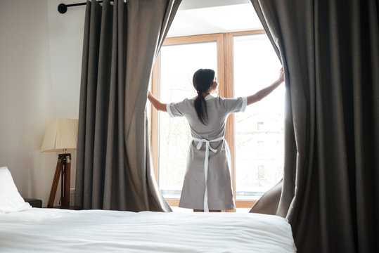 Female chambermaid opening window curtains in the hotel room