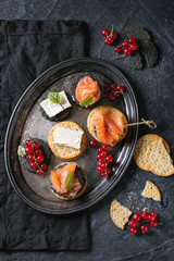 Stack of black charcoal and traditional crackers with smoked salmon, cream cheese, green salad and red currant berries on vintage metal tray over black stone background. Appetizer snack. Top view