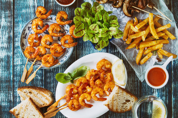 Variety of BBQ snack lunch. Plates grilled spicy king prawn kebabs, mushrooms skewers, bread, french fries potatoes with sauces and greens over blue wooden background. Flat lay