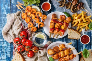 Gardinen Variety of BBQ snack lunch. Plates grilled spicy prawn kebabs, chicken, pork, vegetables, mushrooms skewers, bread, french fries potatoes with sauces and greens over blue wooden background. Flat lay © Natasha Breen