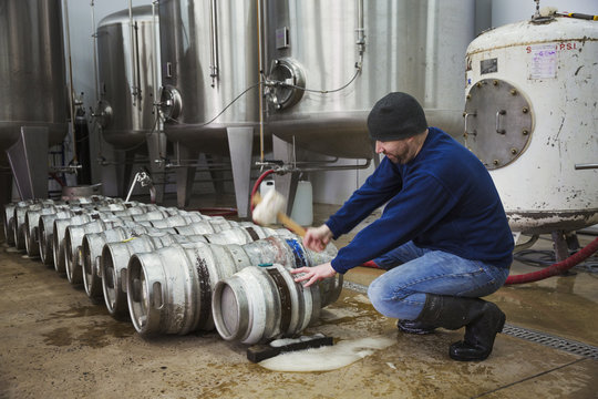 Man kneeling and hammering in a peg into a metal beer keg. Large fermentation tanks. - Powered by Adobe