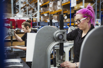A young woman with pink hair, a female factory worker using a hole punching machine, working to assemble a bicycle in a factory.