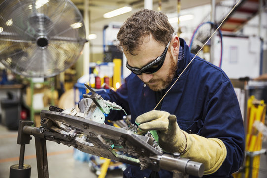 Male skilled factory worker wearing eye protectors welding a bicycle part in a factory.