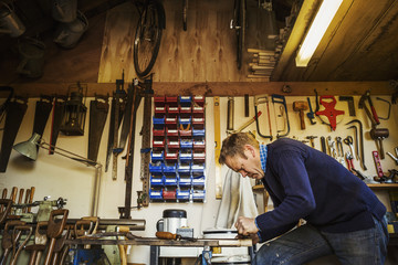 Man standing in a garden workshop surrounded by tools hanging from beams and a stack of garden tools propped up.