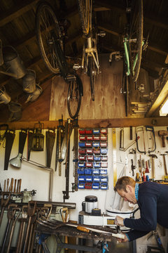 A Man Standing In A Garden Workshop, Working On An Old Garden Space, Surrounded By Tools. Equipment On The Beams And Bicycles Hanging From The Ceiling. 