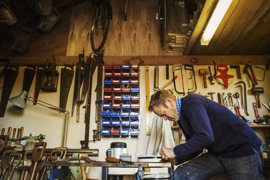 Man standing in a garden workshop surrounded by tools hanging from beams and a stack of garden tools propped up.