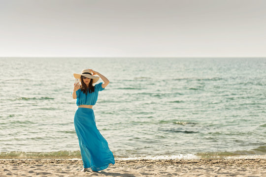 Young beautiful woman on the beach in azure long dress takes pictures of herself on a smartphone.