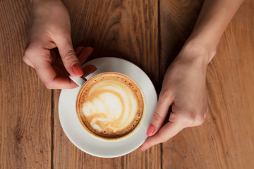 Close-up female hands holding cup with coffee cappuccino with foam with nice pattern. Perfect red gel polish manicure. Wood natural vintage table. Creative color warm post processing