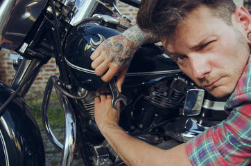 Tattooed man making funny face, while repairing motorcycle, closeup, holding wrench in hand. Selective focus on hand, face slightly out of focus