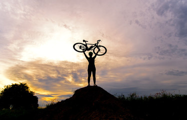 Silhouette the women in action lifting bicycle above his head stand on meadow with sunset