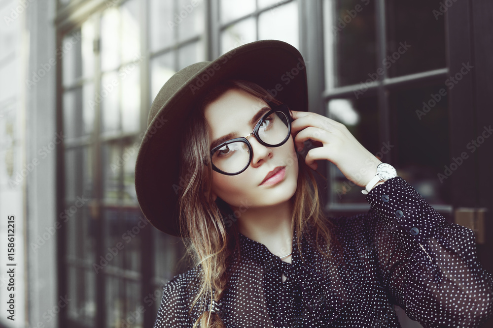 Wall mural Outdoor close up portrait of young beautiful fashionable girl posing in street. Model wearing trendy cat eye glasses, fedora hat, white wrist watch. Female fashion concept. Copy, empty space for text