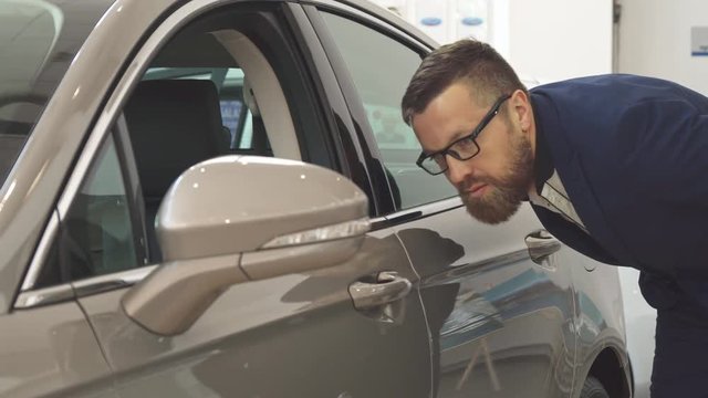 Attractive caucasian man examining new car at the dealership. Bearded male customer touching the door of the vehicle. Handsome mature guy in glasses bending near the gray sedan
