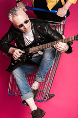 Handsome senior man in leather jacket and sunglasses sitting in shopping trolley and playing electric guitar