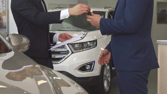 Close up of sales manager in black suit selling the car to the customer. Low shot of seller giving key to the client and shaking his hand. Two men standing at the showroom against background of white