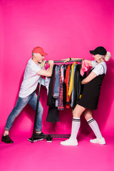 stylish senior couple pulling wardrobe with diferent clothes on hangers isolated on pink, relationship difficulties concept