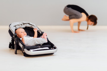 Naklejka premium Little baby lying in child pram while his mother workout on the background. Focus on baby boy. Fitness maternity healthy lifestyle concept.
