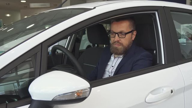 Mature male customer examining car interior at the dealership. Attractive caucasian man touching steering wheel of the vehicle. Handsome bearded guy in glasses sitting inside the car