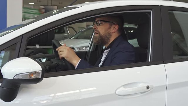 Handsome male customer showing key through the car window at the dealeship. Mature caucasian man sitting on the driving seat inside the vehicle. Attractive bearded guy turning his face to the camera