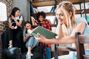 caucasian girl reading book while her friends sitting on sofa at home library