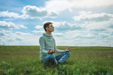 Naklejka na ściany i meble Man traveler with backpack sits on meadow with green grass under blue sky with clouds in the Lotus position