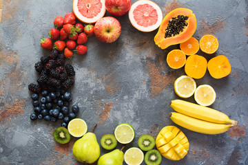 Rainbow color fruits arranged in a circle, strawberries, blueberries, mango, orange, grapefruit, banana, apple, grapes, kiwis, papaya on the grey background, copy space for text, selective focus