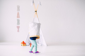 Funny little kid running around the bright room with a paper bag on head on the background of children's wigwam © gorynvd