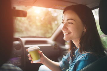 Beautiful woman smiling while sitting on the front passenger seats in the car and she is drinking coffee from a disposable cup