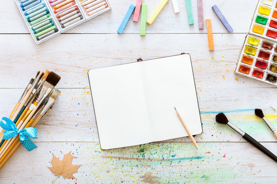 Artistic design workspace, notebook mock up for artwork with watercolor paints, pencil, set of different paintbrushes on white wooden table, back to school, children kid art. Top view and copy space