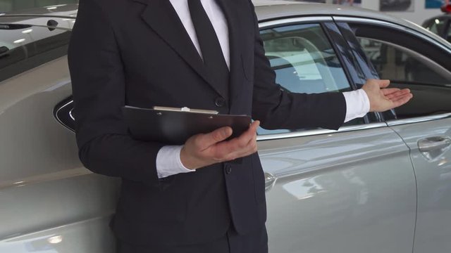 Close up of sales manager pointing his hand on the new car at the dealership. Low shot of male seller in black suit holding clipboard in his hands. Consultant offering to look at the luxury gray