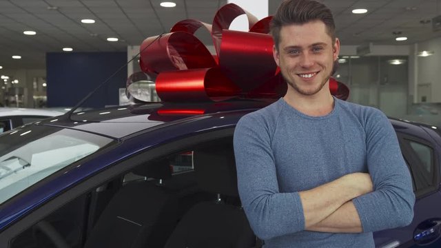 Handsome male customer posing near the new car at the showroom. Attractive young man showing key near the purple hatchback. Caucasian guy folding his arms on his chest against background of vehicle