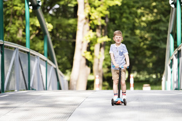 Boy riding his scooter in the local park. Sunny summer.