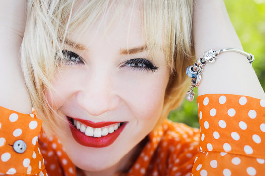close-up portrait outdoors young beautiful girl in an orange hipster blonde bright cheerful polka dot blouse , smiling red plump lips on the background of green grass