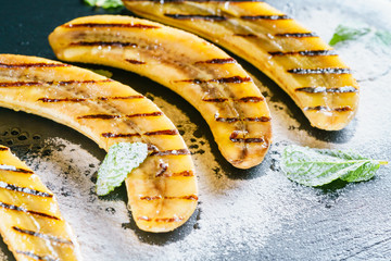 Halves of grilled bananas are nicely laid on a black kitchen board and shale stone. Dish is poured with clear honey, sprinkled with sugar powder and decorated with green mint leaves. Close Up.
