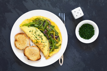 Omelette with green bell pepper and red onion sprinkled with chives, served on plate with toasted and buttered bread slices, photographed overhead on slate with natural light