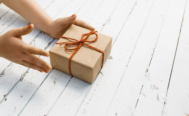 Craft paper gift box with red bow in the children's hands on white wood background.