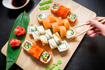Sushi rolls on wood board, sauce, ginger, wasabi and hand holding chopsticks on dark background. Top view. Flat lay.