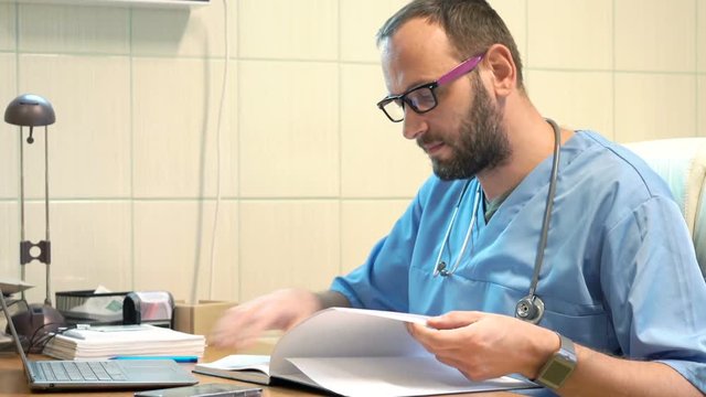 Young, male doctor working with documents and laptop sitting by table in office
