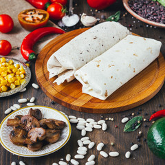 Vegetarian burrito on wooden board over black table surrounded by ingredients.