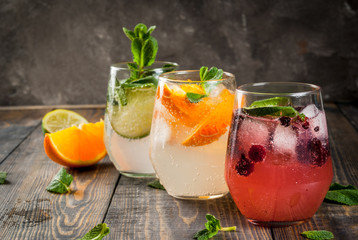 Selection of three kinds of gin tonic: with blackberries, with orange, with lime and mint leaves. In glasses on a rustic wooden background. Copy space