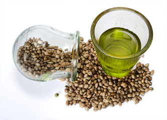 Hemp seeds in a round glass jar and hempseed oil in a measuring glass beaker on a white background.