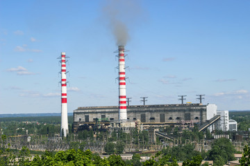 Pipe industrial plant with smoke against bright blue sky and above green trees