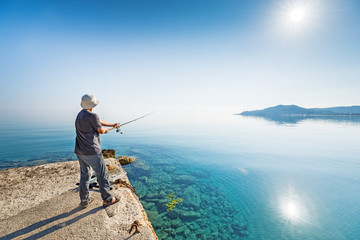 Side view of young man fisherman standing on pier with rod. Seashore of Ionian sea, Zante - Zakinthos island, Greece. Fishing background. Sunrise morning scenery.
