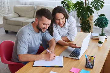 Man writing in notebook while woman showing something to him in laptop