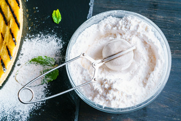 Metallic kitchen strainer lies in a glass bowl with powdered sugar, standing on a black table top near shale stone plate with grilled banana dessert. Macro Closeup Top View.