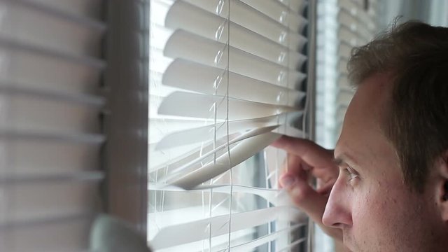 A guy with pimples on his face looking through the blinds in the window
