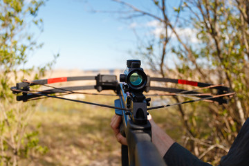 A crossbow with a sight to aim in first person on the background of the lake. The man preys on wild animals in summer forest