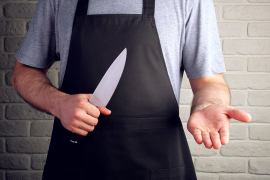 man in black apron holds knife in his hands open palm, on background of brick wall
