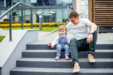 A young father and a little daughter are resting on the steps in the city park.
